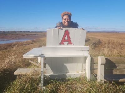 Angel Percentie smiles and poses on an outdoor chair painted with the Acadia "A" on a cold day on Wolfville's dylelands and harbour.