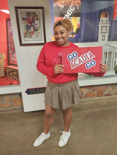 Angel Percentie poses in the Acadia Athletics Complex while wearing a red Acadia sweatshirt and holding a "Go Acadia Go" sign