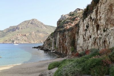 Beautiful Greek coastline: rocky cliffs with old stone buildings atop them and a bright beach with blue water.