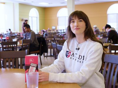Dietitian Paula Farrell ('20) smiles while sitting at a table in Wheelock Dining Hall wearing a white Acadia University sweatshirt.