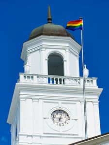 The rainbow pride flag flutters in the wind in on a flagpole in front of the Acadia clock tower.