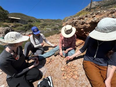 Students and faculty in sun hats sift through a pile of shards and rock samples in a rocky Greek landscape