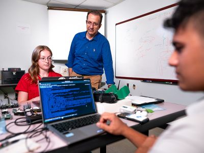 Dr. Esteve Hassan observes as two students in a computer electronics lab read code on laptops.