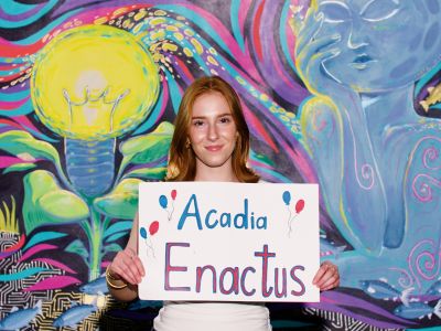 Sophia smiles in front of a painted mural while holding an "Acadia Enactus" sign.