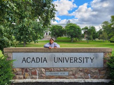 Angel Percentie smiles and poses with the stone "Acadia University" sign, with University Hall visible in the background.