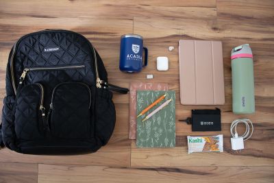 An overhead view of a backpack and its contents, including a mug, notebooks, tablet, pouch, granola bar, USB charger, and water bottle.