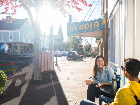 Two students laugh and chat over coffee in the middle of downtown Wolfville, with bright sunlight striking the neon "Acadia" sign.