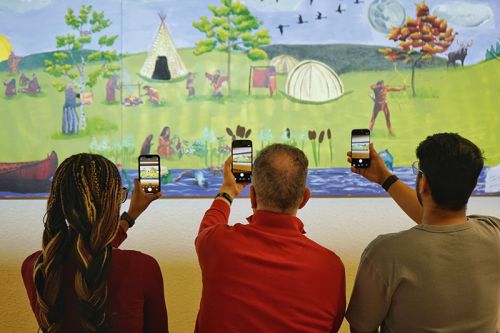 Joyce, Esteve, and Nitish each point their smartphone apps at a large painting of Indigenous peoples in a museum.