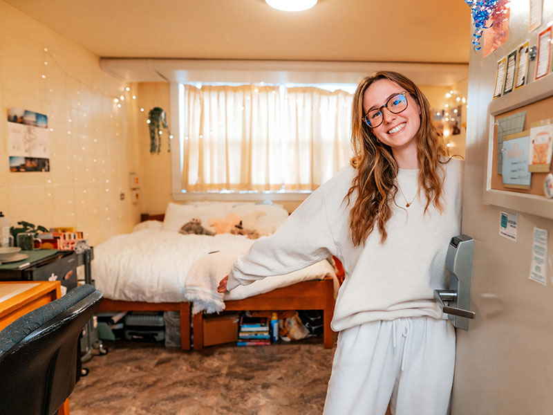 A female student cheerfully welcomes the viewer to a residence room