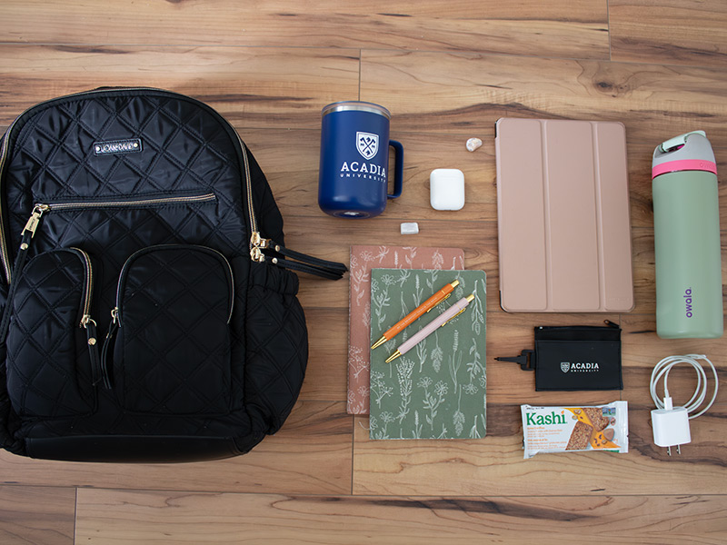 An overhead view of a backpack and its contents, including a mug, notebooks, tablet, pouch, granola bar, USB charger, and water bottle.