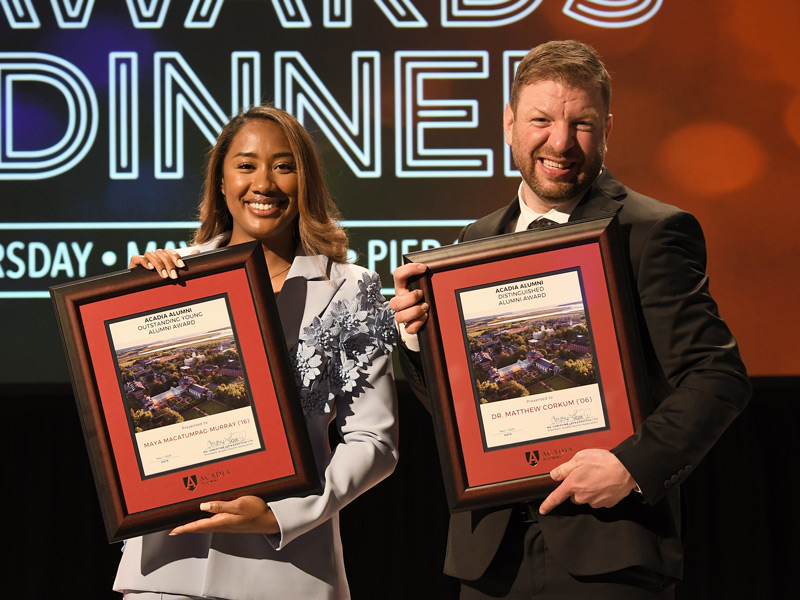 Maya Macatumpag-Murray and Dr. Matthew Corkum smile as they stand on stage with their alumni award plaques at the annual Alumni Dinner event.