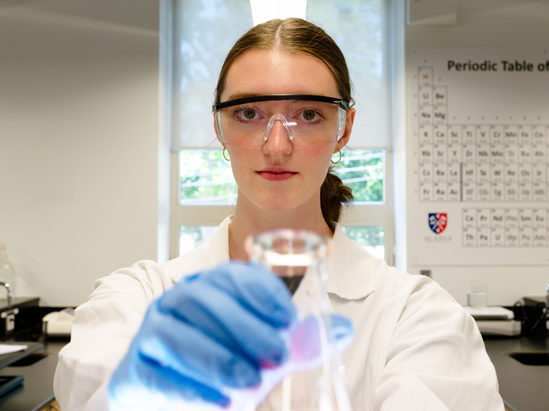 Anna Lukeman holds a laboratory beaker in a chemistry laboratory and looks at the camera.