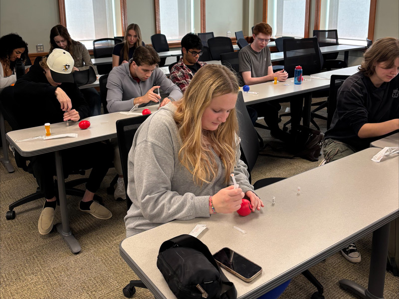 A group of students practice administering Naloxone using training kits in a Patterson Hall classroom.