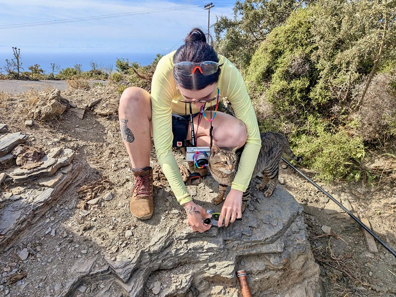 A female Acadia student kneels down atop a rock to examine a specimen while a cat curls up to her.