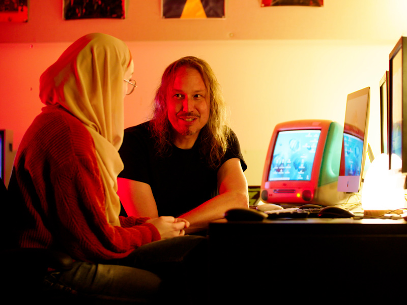 Professor Jon Saklofske and a student in discussion, seated next to vintage Apple and PC computers.