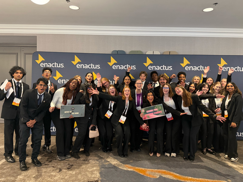 Over a dozen Enactus Acadia students in business attire cheer in front of an enactus banner in a conference venue lobby.
