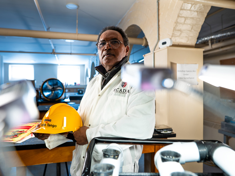 Dr. Martin Tango is pictured between stacks of equipment in an engineering workshop space at Acadia. He wears a lab coat and holds a hardhat.