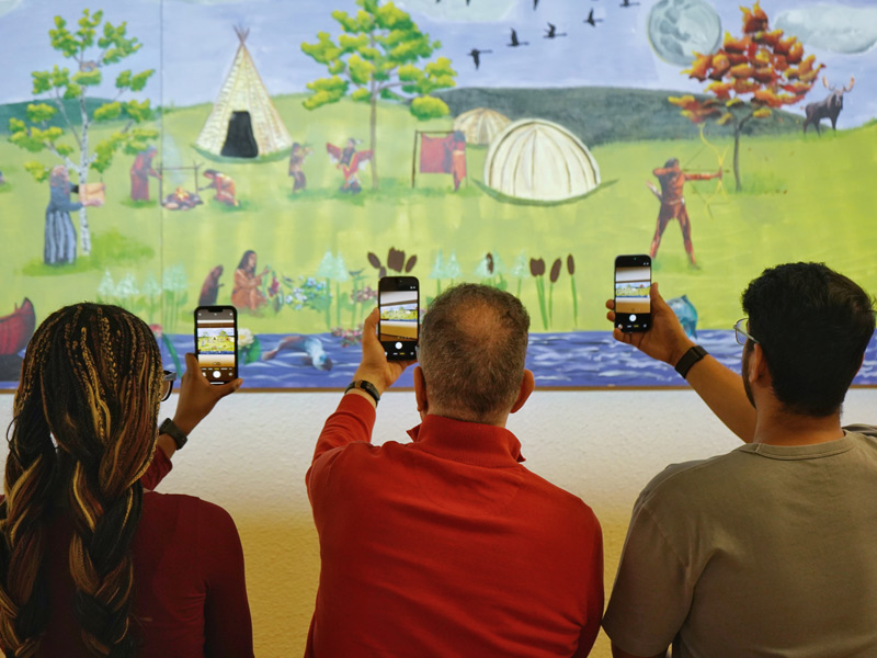 Joyce, Esteve, and Nitish each point their smartphone apps at a large painting of Indigenous peoples in a museum.