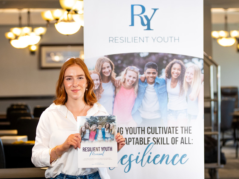 Sophia smiles while holding up the Resilient Youth Manual and standing in front of Resilient Youth signage in a conference room.