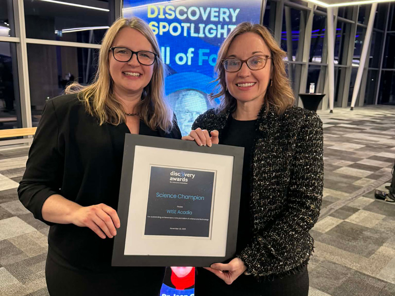 Drs. Melanie Coombs and Randy Newman smile together while holding a plaque reading "Discovery Awards- science champion" in a conference centre setting.