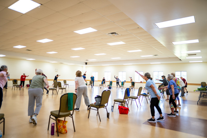 A group of seniors standing behind chairs arranged in a circle, stretching one arm forward.