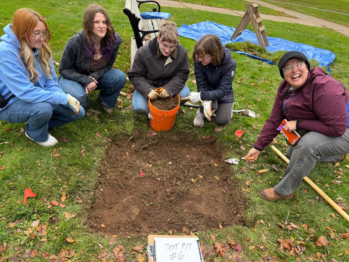 A group of students smile around a patch of removed turf labelled "Test Pit 6" on the University Hall South Lawn.