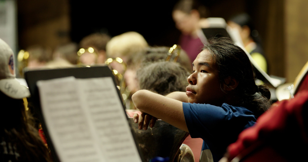 A student leans on their brass instrument during a rest in a concert band rehearsal.