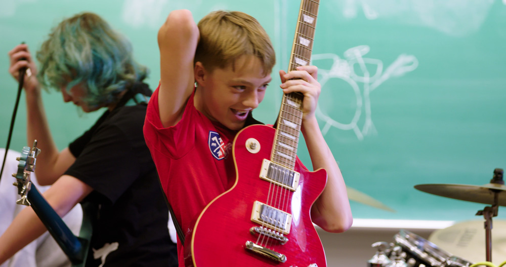 A student in a red Acadia shirt smiles as he lifts up a red electric guitar.