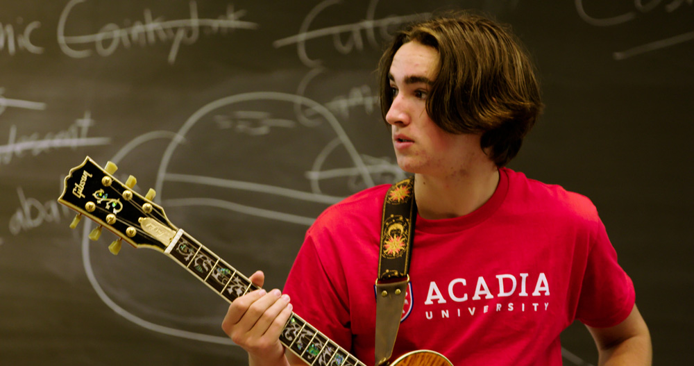 A teenage band camp student holding an acoustic guitar looks towards an instructor.