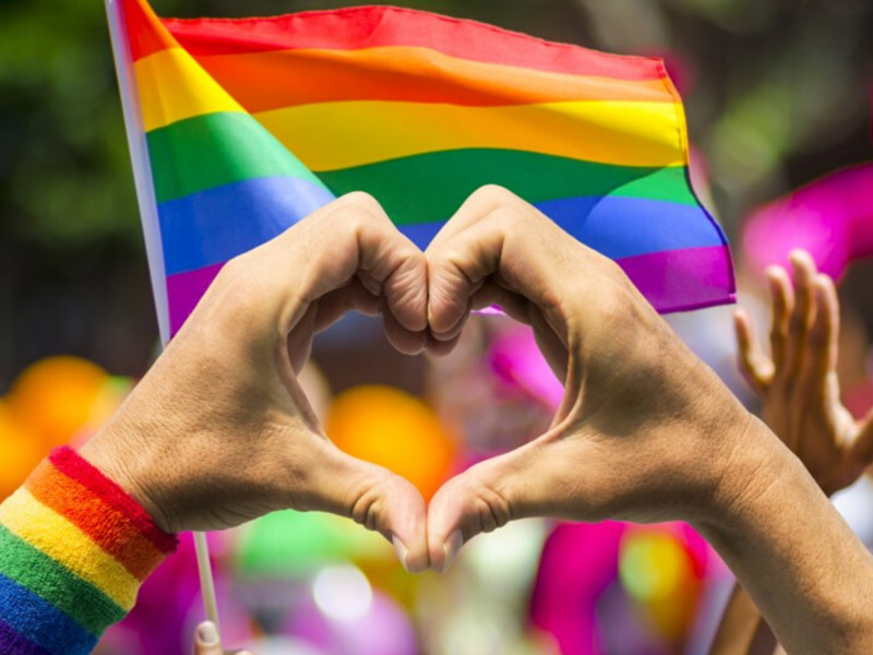 Hands forming a heart in front of a rainbow Pride flag.