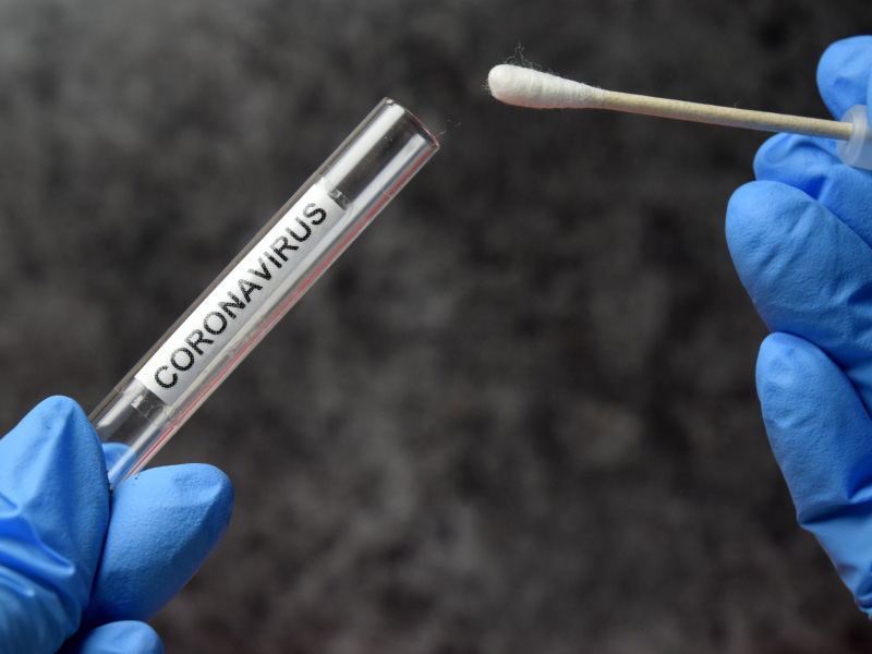 Gloved hands hold a sample vial labelled "Coronavirus" and a testing swab
