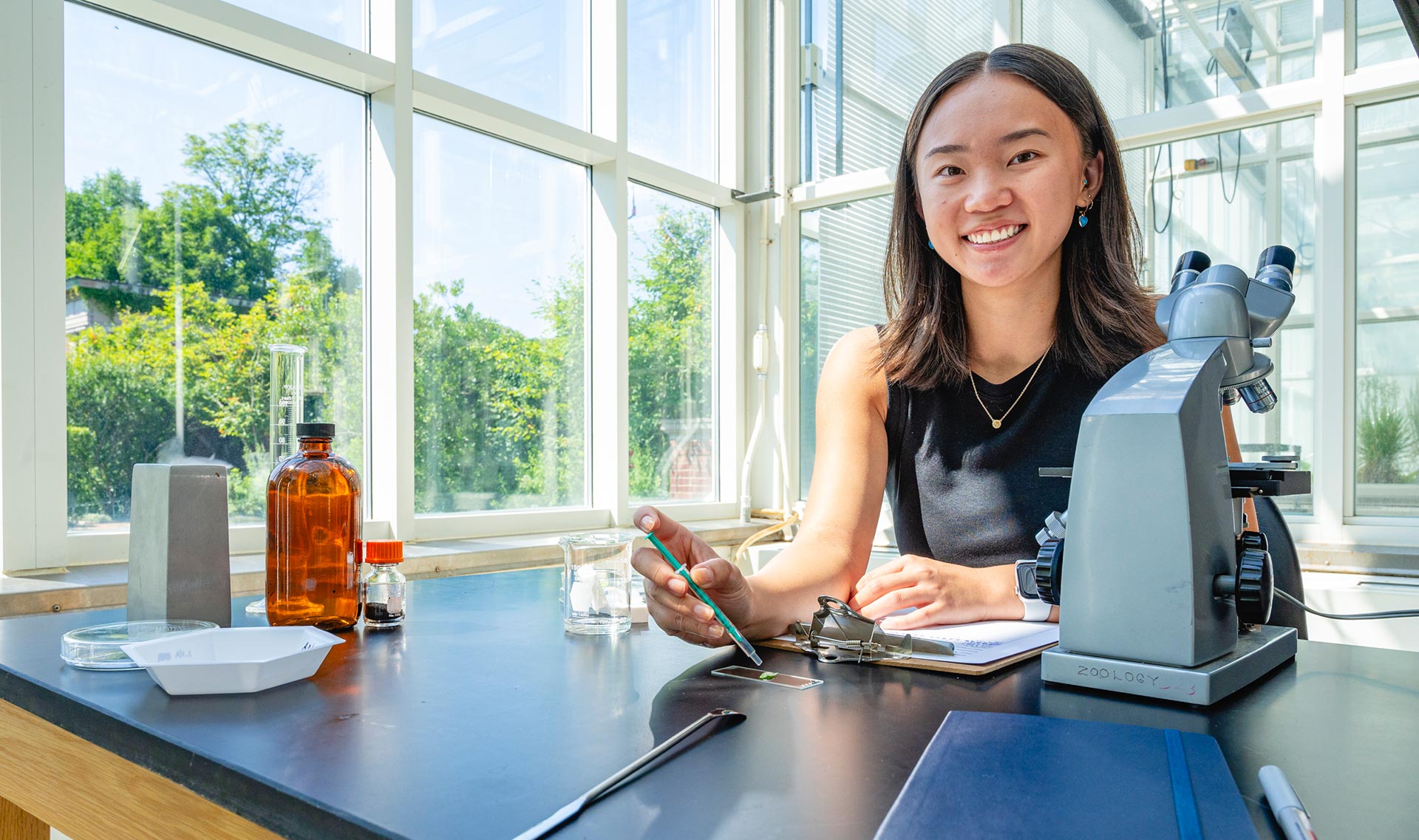 A female biology student smiles as she works with a microscope and sample slide in an airy lab space.