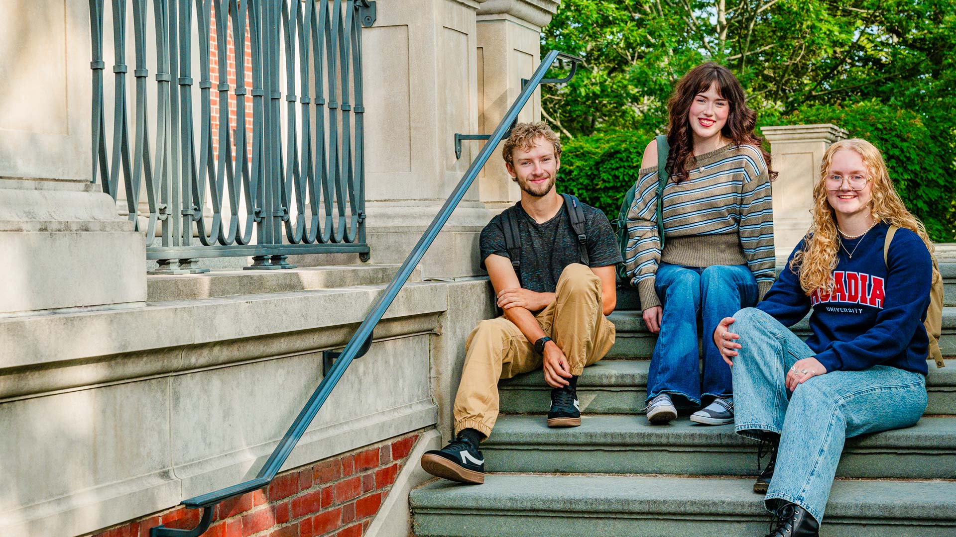 Three students smile as they sit casually on the stone steps of the KC Irving Environmental Science Centre, with. bright green ivy and trees behind them.
