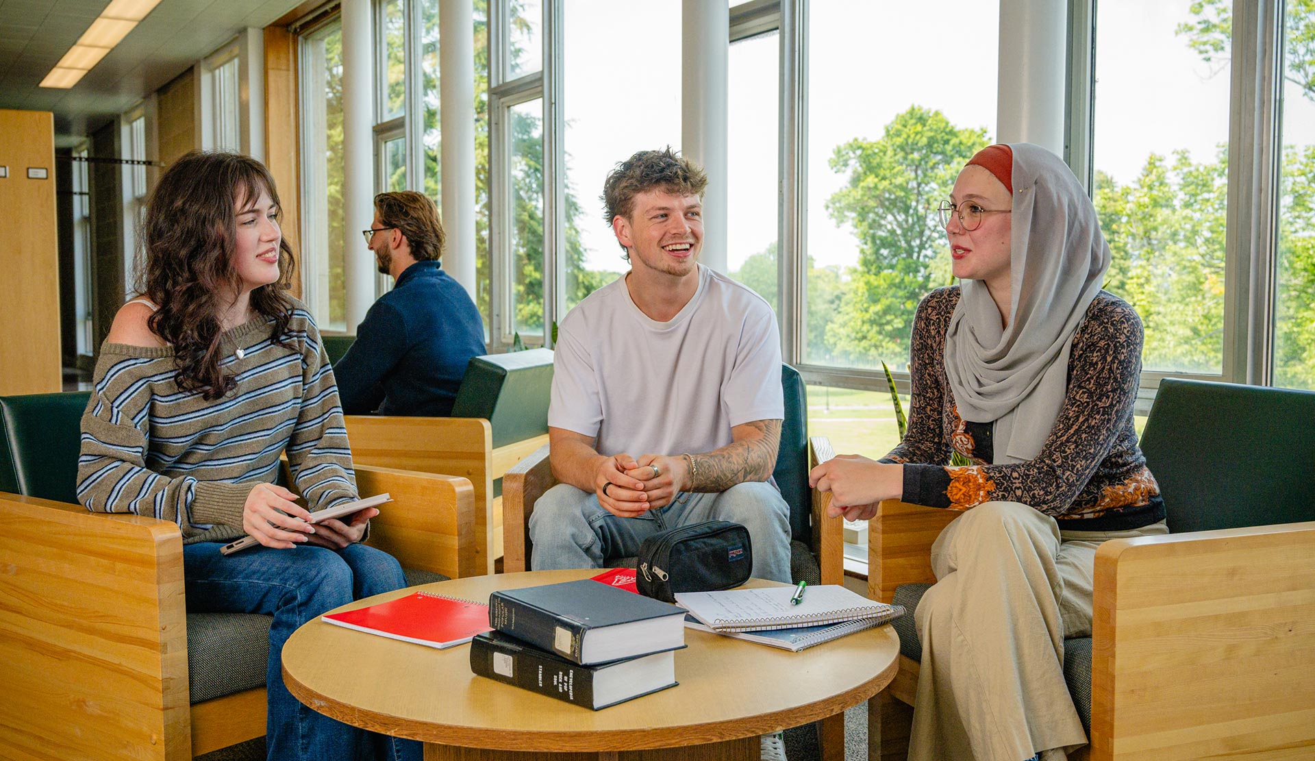 Three students chat in chairs in the Vaugahn Memorial Library, with books and study materials in front of them. Outside the large windows, it is a bright day.